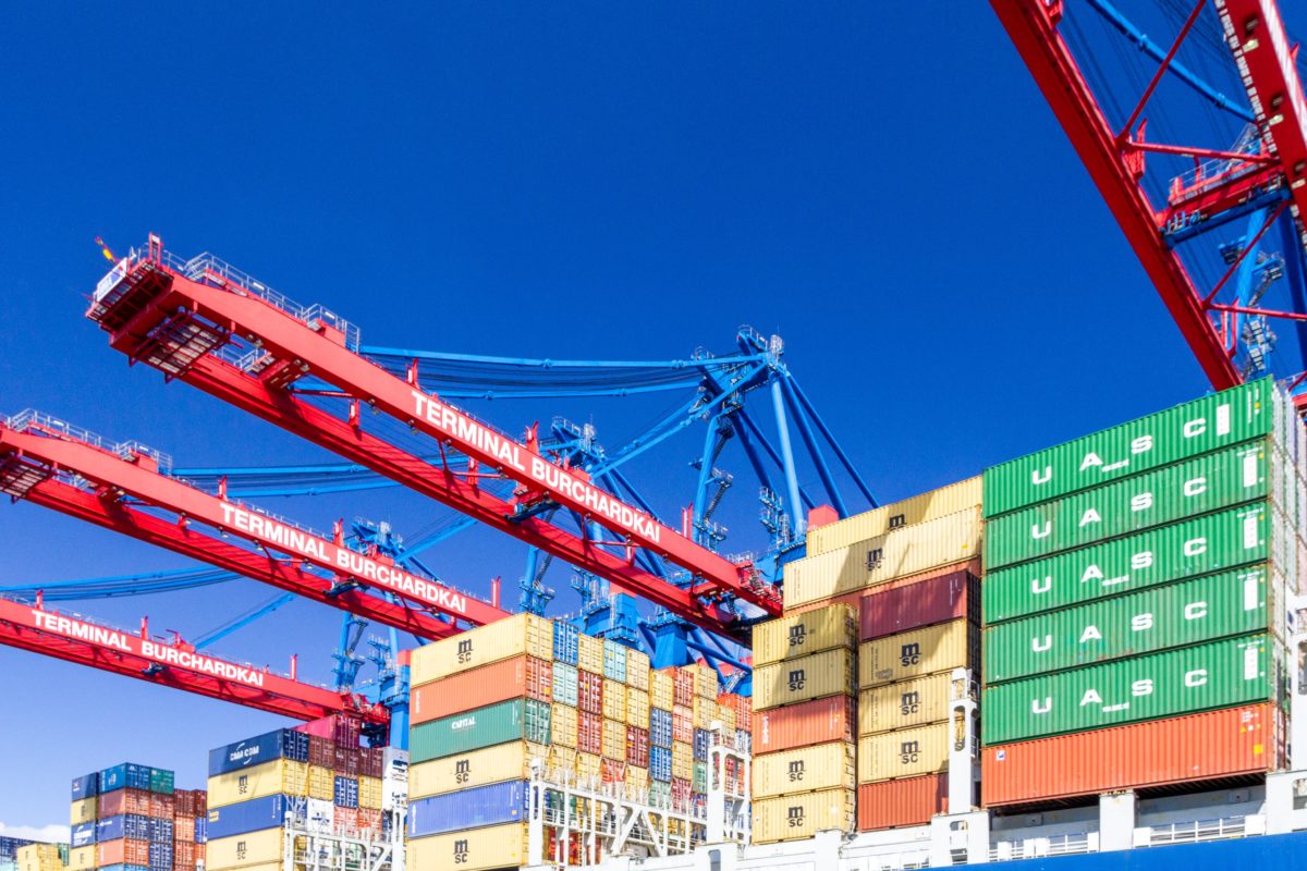 Shipping containers with red cranes overhead, underneath a clear blue sky
